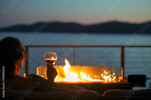 A woman sitting in an elegant setting at night outside drinking a glass of red wine with a warm fire overlooking a lake view. 