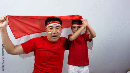 Asian father and son with happy and cheerful expressions wearing red tops and headbands while holding Indonesian flags, isolated on white background. Indonesian Independence Day concept.