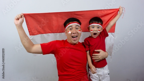 Asian father and son with happy and cheerful expressions wearing red tops and headbands while holding Indonesian flags, isolated on white background. Indonesian Independence Day concept.