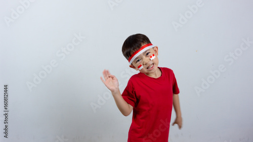 A cheerful Indonesian boy wearing a red top and headband, a mini Indonesian flag sticker on his face, posing hello and smiling, isolated on a white background. Indonesian Independence Day concept.