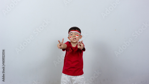 Happy Indonesian boy wearing red top and headband, miniature Indonesian flag sticker on his face, posing peacefully with his finger, isolated on white background. Indonesian Independence Day concept