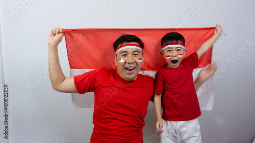 Asian father and son with happy and cheerful expressions wearing red tops and headbands while holding Indonesian flags, isolated on white background. Indonesian Independence Day concept.