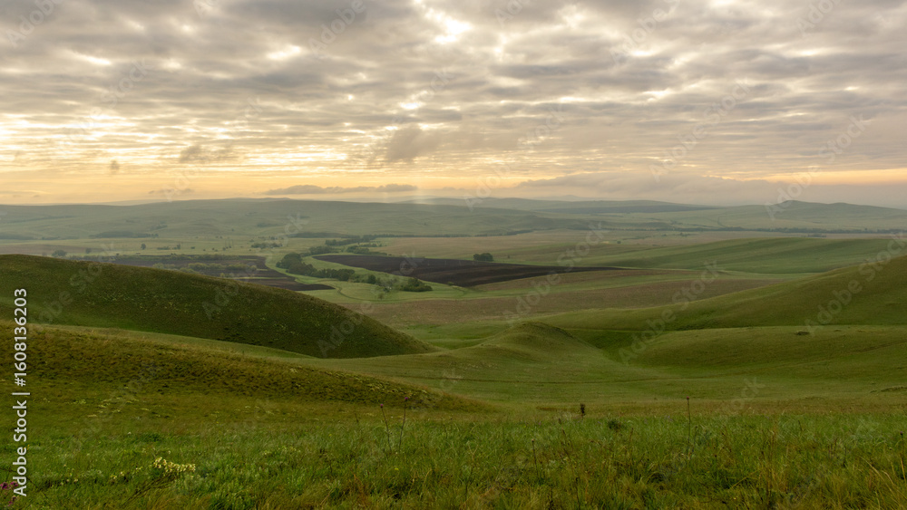 Fototapeta premium Expansive green hills under a cloudy sky at dawn, showcasing rolling landscapes and distant fields, creating a serene and tranquil atmosphere in nature's beauty