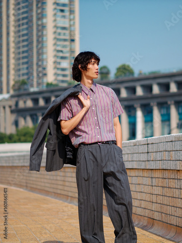 Portrait of handsome Chinese young man with black curly hair wearing stripe shirt posing with suit on shoulder with urban background. Male fashion, cool Asian man lifestyle.