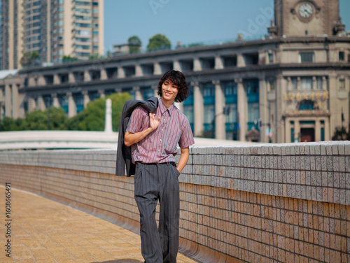 Portrait of handsome Chinese young man with black curly hair wearing stripe shirt posing with suit on shoulder with urban background. Male fashion, cool Asian man lifestyle.