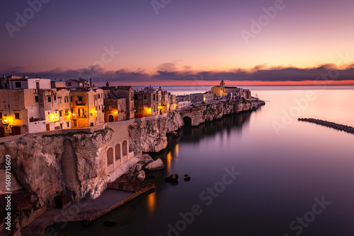 Fototapeta Naklejka Na Ścianę i Meble -  Coastal town of Vieste, Italy, captured during twilight using long exposure technique, highlighting warm street lights, dramatic cliffs, and a serene Adriatic Sea.