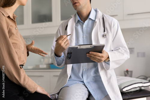 Young general practitioner man giving consultation in clinic office, talking to patient woman on examination couch, holding clipboard, writing notes for medical history. Cropped shot