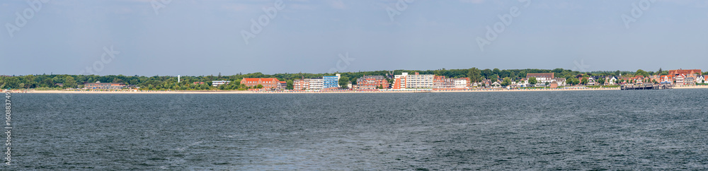 Fototapeta premium Panoramafoto von Wyk auf Föhr bei schönem, sonnigen Wetter mit Strandpromenade, Hafen und Gebäuden vom Meer aus gesehen mit dem Wasser der Nordsee im Vordergrund
