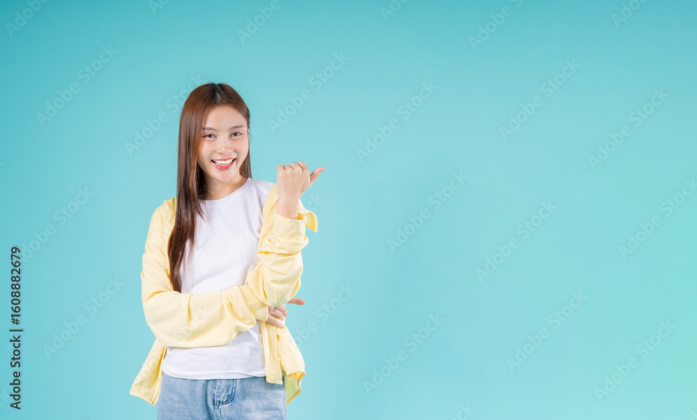 Cheerful young Asian woman pointing to the side while smiling against a turquoise background, representing advertisement, presentation, promotion, and positive lifestyle concepts. Cheerful young Asian woman pointing to the side while smiling against a turquoise background, representing advertisement, presentation, promotion, and positive lifestyle concepts.