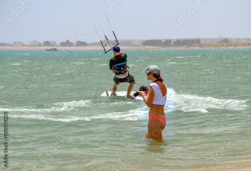 Young woman photographing kitesurfers on a sunny beach in Hurghada, Egypt. Vibrant seaside moment capturing action, wind, waves and coastal energy of the Red Sea.
