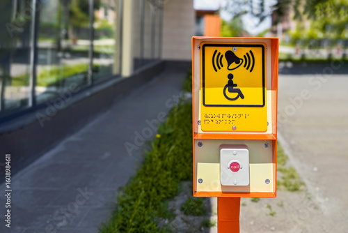 Orange accessibility button with yellow sign stands on city sidewalk. Sign wheelchair symbol, bell icon, and Braille text, aiding disabled individuals. Modern building frame the scene under daylight.
