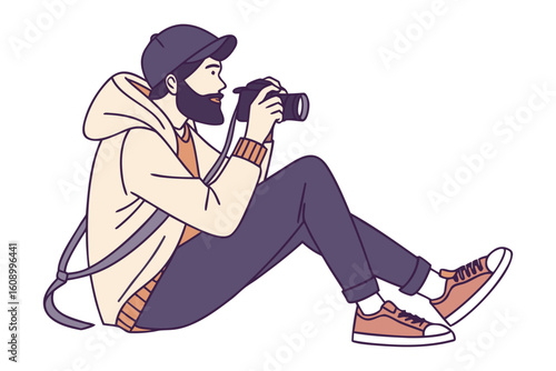 A stylishly dressed bearded man wearing a baseball cap and casual clothing sits on the ground taking a photograph with a digital camera