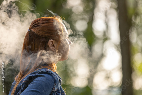 An young woman with long, red hair vaping in a forested area. The person is captured from the side, exhaling a thick cloud of vapor from an electronic cigarette. Summer landscape in a background.