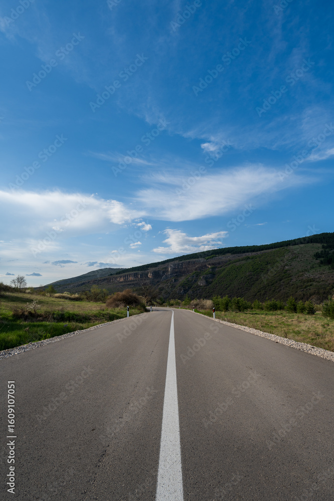 Fototapeta premium An empty highway that passes through fields and goes towards the mountain. The road is covered with blue sky and beautiful fluffy clouds. 