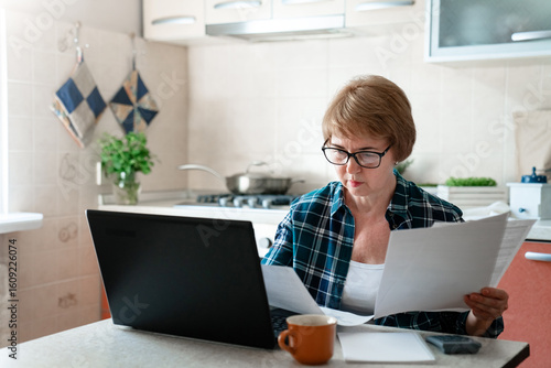 Tableau sur toile Senior woman working from home kitchen with laptop, documents, finances