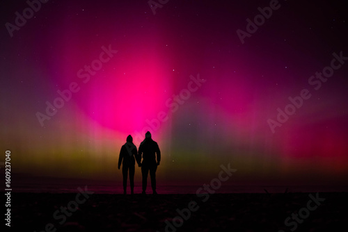 Silhouette of couple holding hands and gazing at sky lit up by vibrant pink and green southern lights, Aurora australis, on beach near Christchurch, Canterbury, South Island, New Zealand