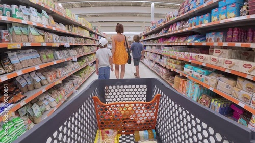 Family shopping in supermarket, viewed from shopping cart perspective. Mother with two kids walking down aisle filled with food products and groceries