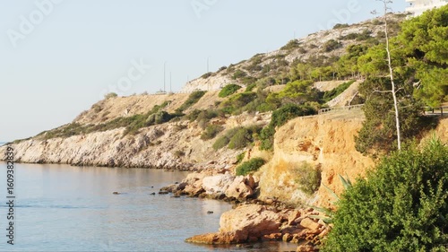 Wallpaper Mural Rocky seashore with calm water, light vegetation, and steep cliffs in Varkiza, Athens. Hill in background with winding road and streetlights. Lower pedestrian path with wooden railing.  Torontodigital.ca