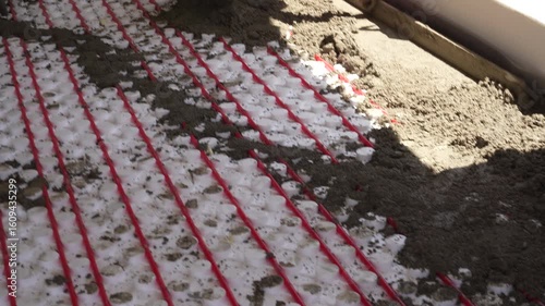 Workers pour concrete mortar onto the top of underfloor heating pipes during construction.