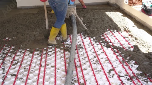 Workers pour concrete mortar onto the top of underfloor heating pipes during construction.