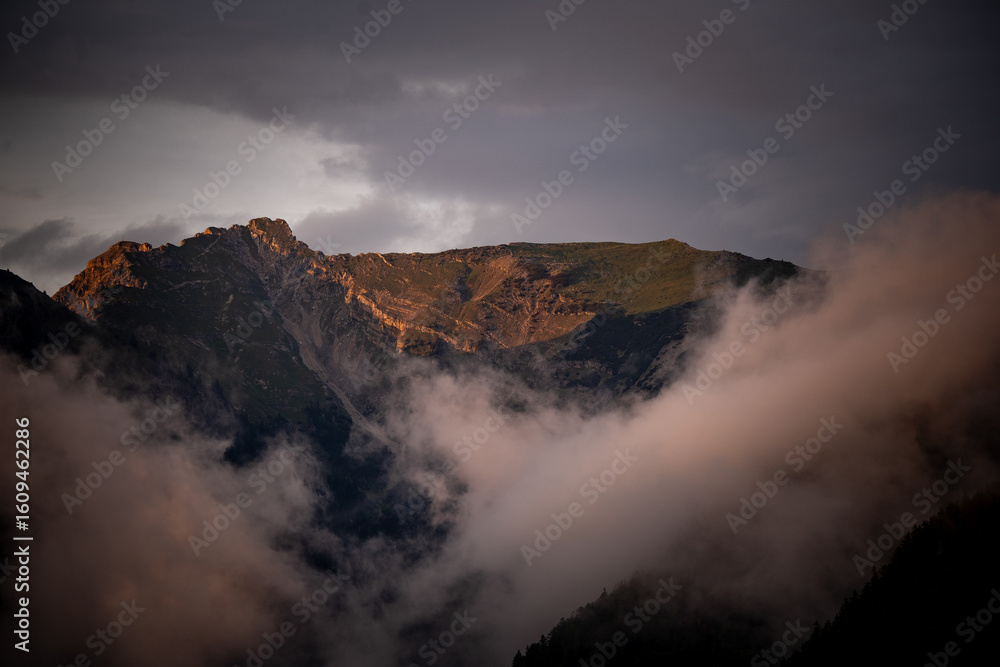 Obraz premium Dramatic Alpine Sunset: Golden Light on Rugged Peaks & Misty Clouds – Majestic Mountain Landscape