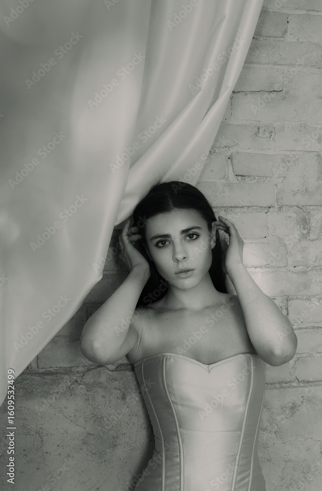 Fototapeta premium A young woman wearing a white corset dress standing against a textured wall with hands near her head