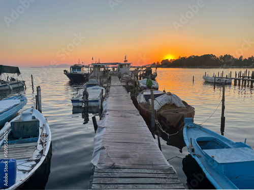A peaceful sunset at the marina with boats gently floating on calm waters. The wooden pier leads the eye to the horizon where the sun sets, painting the sky in warm orange and purple hues.