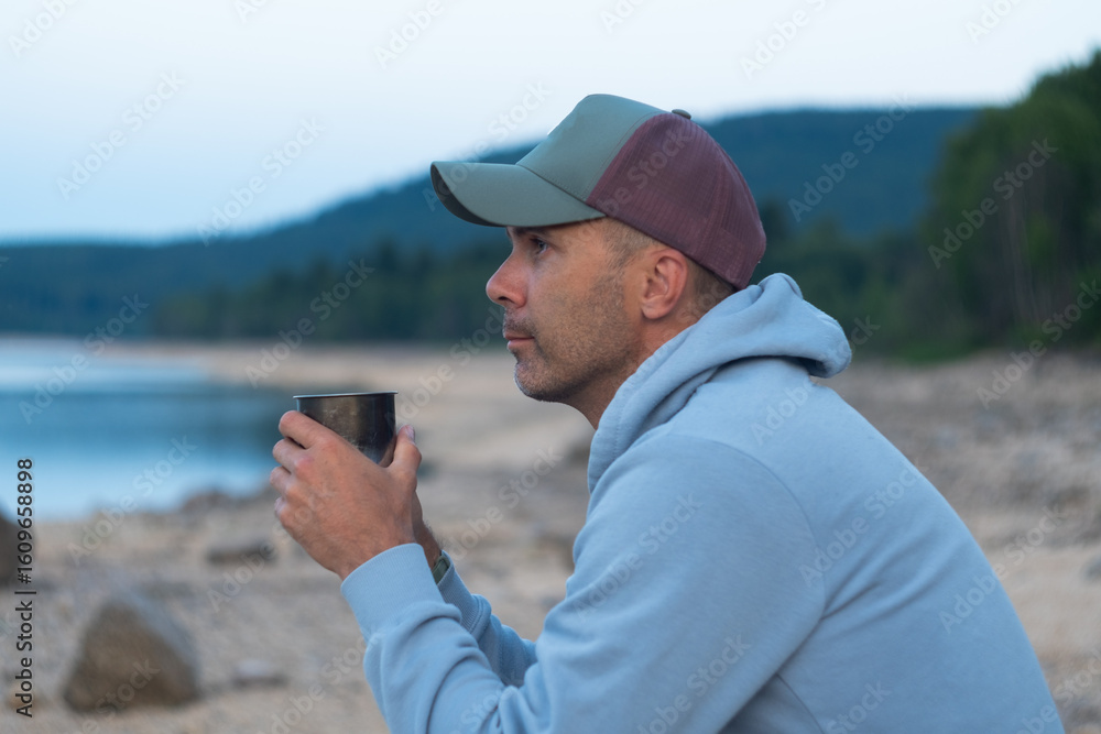 hiker man in hat is sitting on lake coast with mug of coffee. travel concept hiker man in hat is sitting on lake coast with mug of coffee. travel concept