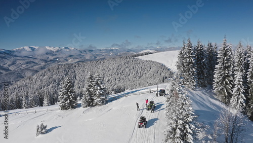Aerial drone footage tourists riding snowmobiles and taking pictures in a snowy mountain forest, enjoying a breathtaking winter landscape on a sunny day. Bukovel ski resort. Winter travel background