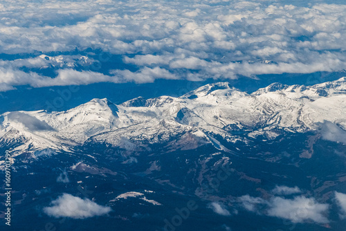 Aerial view of snow-capped peaks in the Grand Teton range, part of the Rocky Mountains. The scene captures the dramatic mountain tops emerging through a dense layer of clouds