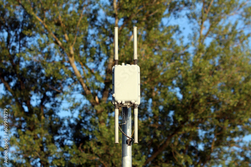 A small, box-like wireless access point with four short antennas is installed atop a metal pole, providing Wi-Fi or cellular signal distribution in a leafy suburban or park environment