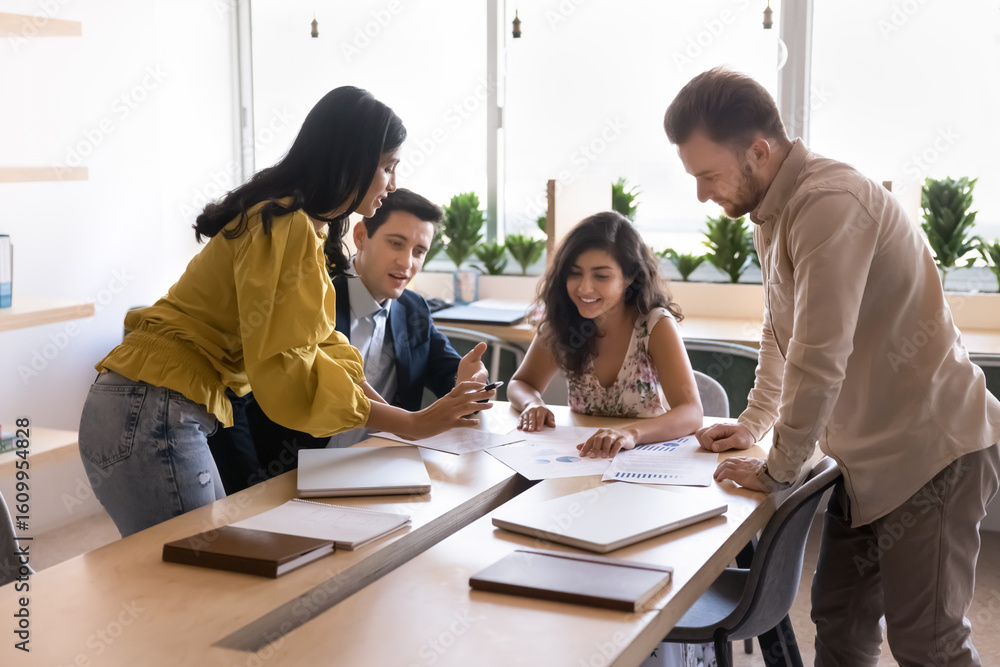 © fizkes - Group of four multiethnic workmates engaged in brainstorming session, reviewing data in documents and charts placed on table, working, thinking and analyzing together during corporate briefing event