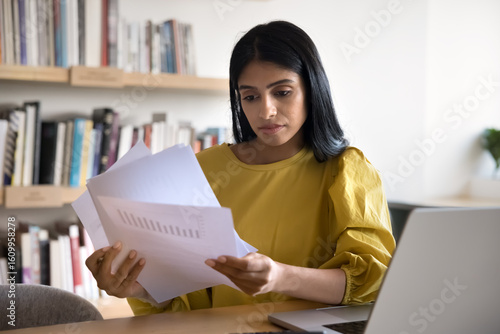 Businesswoman reviewing printed documents while sit at desk with laptop, analyzing content of pages with charts, data, information, examining financial report, project updates, or business documents