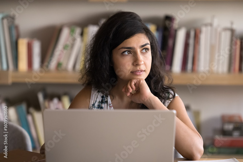 Indian woman working on laptop, looks focused and thoughtful, analyzing or reflecting on information, reviewing report, considering potential decisions or strategies for upcoming project or meeting
