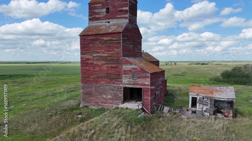 Red grain silo sits in a field with a cloudy sky overhead. The silo is old and abandoned, and the surrounding area is mostly empty. Scene is somewhat melancholic, as the old building