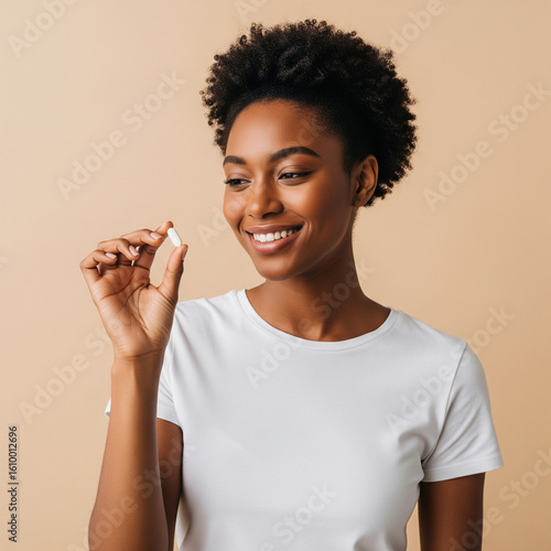 Happy healthy young adult African American woman holding vitamin in hand looking at pill standing isolated on beige background advertising beauty supplements nutrition treatment concept.