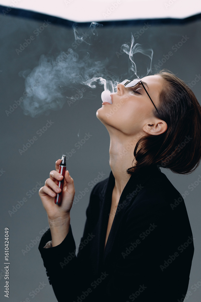 Fototapeta premium Young man vaping and blowing smoke rings with e-cigarette wearing glasses and black jacket