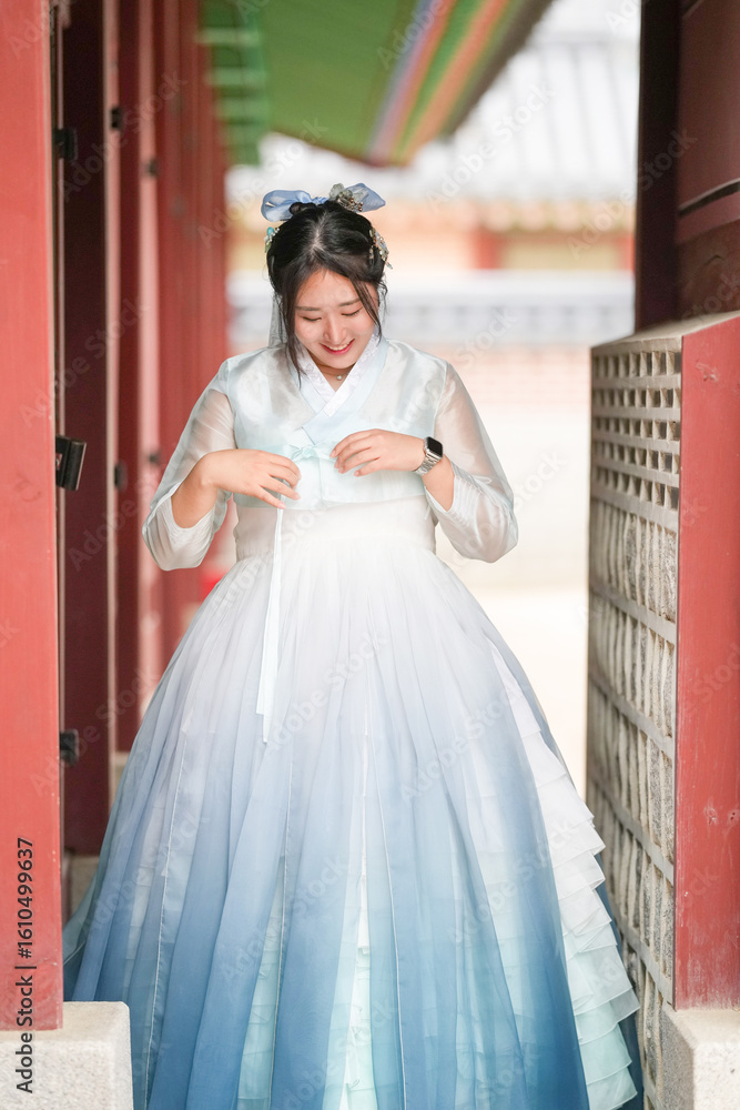 Naklejka premium A Korean woman in her twenties walks through the historic corridor of Gyeongbokgung Palace, wearing a charming sky-blue hanbok under a clear summer sky, framed by ornate wooden pillars. Seoul, June.