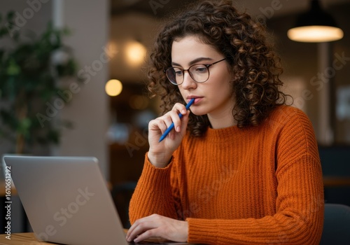 A young woman with curly hair and glasses wearing an orange sweater is focused on her laptop, holding a pen to her mouth in deep thought