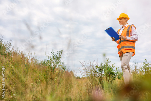 A female engineer wearing a safety vest and yellow helmet communicates via mobile phone while holding a clipboard, standing outdoors in a grassy area