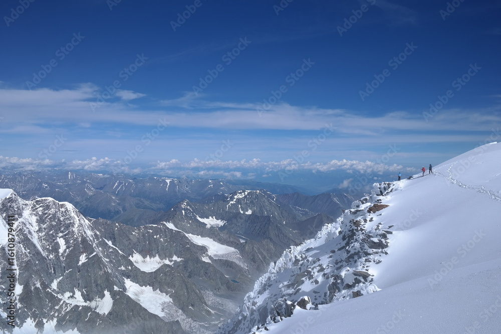 Naklejka premium A lone hiker, warmly dressed in winter gear, stands triumphantly at the summit of a snow-covered mountain, admiring the breathtaking landscape under the clear blue sky and bright sun.