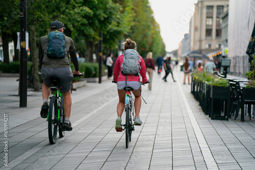 Couple cycling on urban pedestrian street with backpacks, tree-lined walkway, people walking, cafes, and modern city atmosphere in vibrant European downtown lifestyle setting