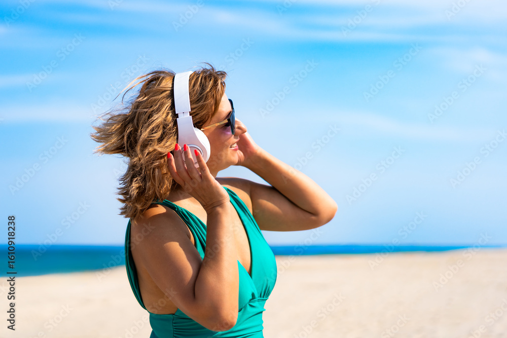 Naklejka premium Summertime with music. Portrait of beautiful mature woman listening to music in headphones on sandy beach