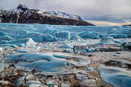 Vast blue glacier with rugged ice formations and distant snowy mountains in Skaftafell, Iceland, under a cloudy sky.