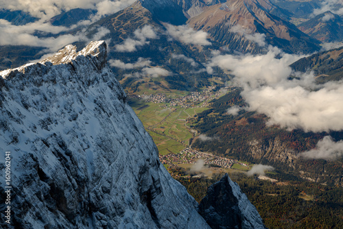 Snow-capped Alpine peak overlooking a vibrant valley village and misty clouds in the Austrian Alps, showcasing dramatic mountain landscape.