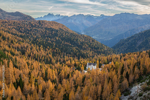Vast autumn larch forests in a Dolomites valley, Italy, with a white building nestled amidst golden trees and distant peaks under a clear sky.