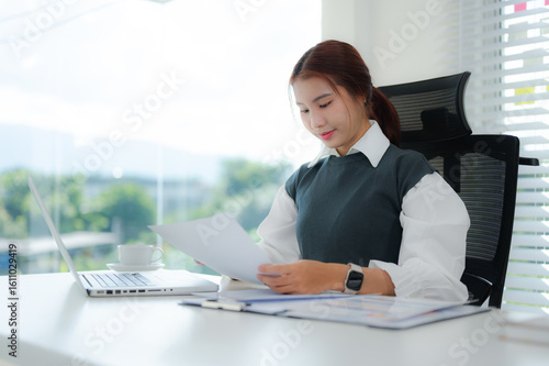 Картината върху платно A diligent young Asian businesswoman is intently reviewing a document while seated at her bright office desk
