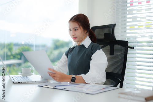Фотография A diligent young Asian businesswoman is intently reviewing a document while seated at her bright office desk