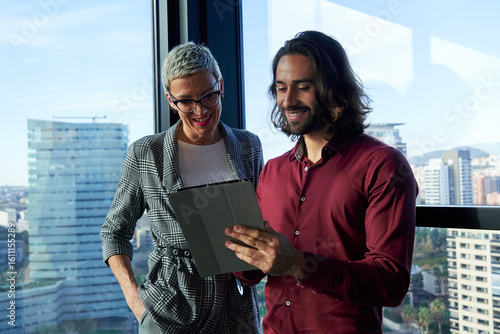 Two smiling businesspeople using a tablet, working together in a modern office with a view of the city skyline.