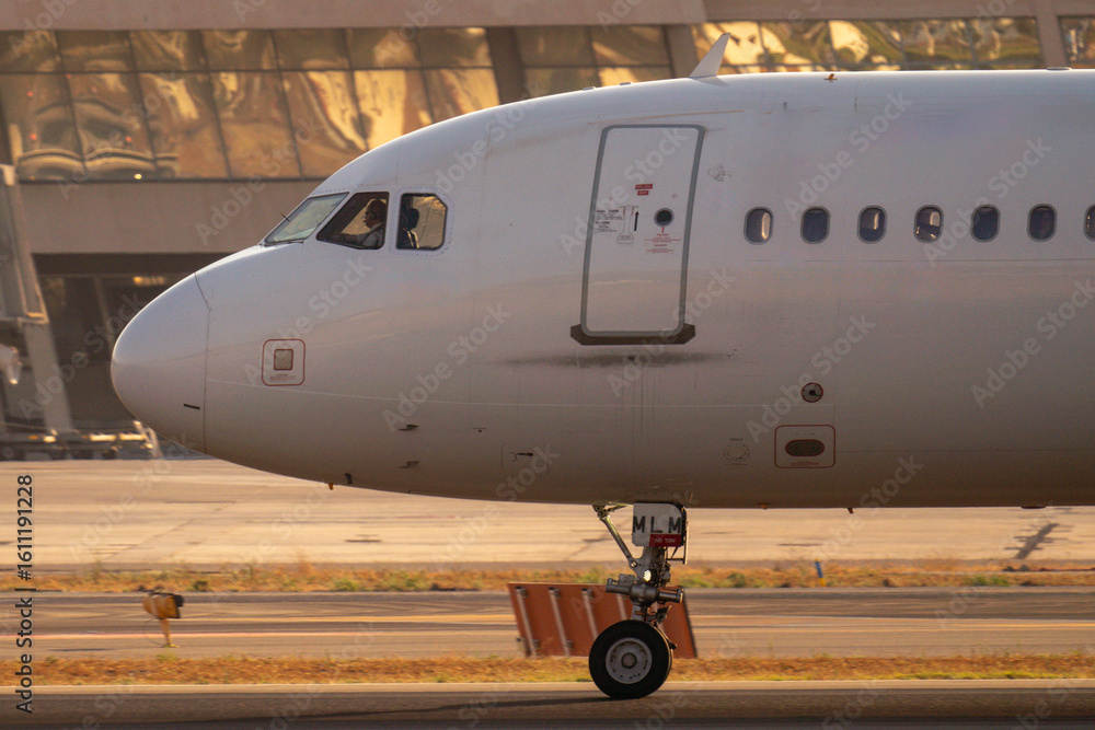 Obraz premium Primer plano del morro de un avión comercial con el piloto visible en cabina rodando por pista frente a la terminal del aeropuerto al atardecer